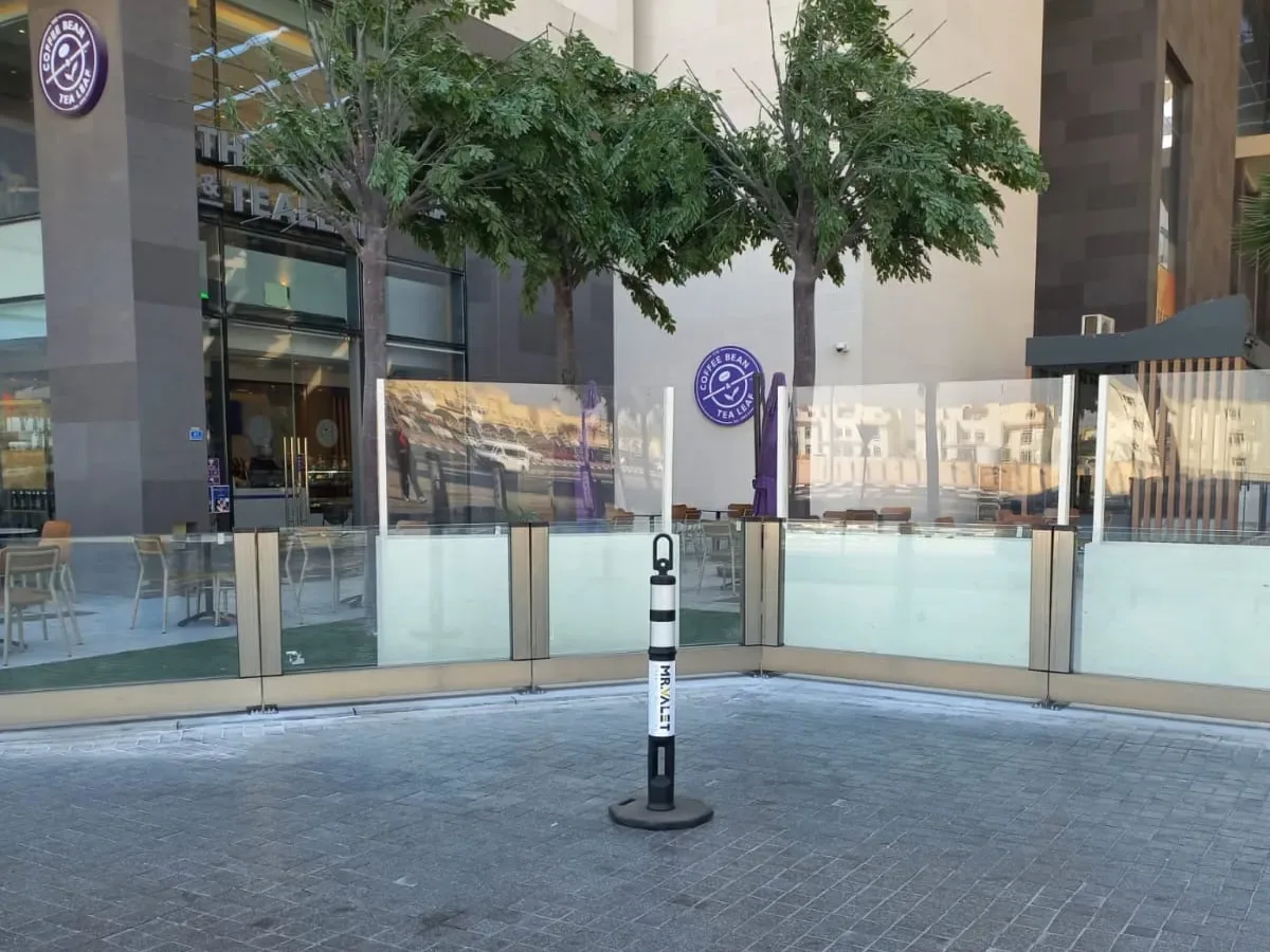 Bir meydandaki kafe terasını çevreleyen cam rüzgâr kırıcı paneller
